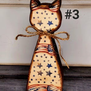 May include: Wooden cat-shaped decoration with a patriotic theme. The cat features a brown outline, with a design of the American flag and blue stars. A twine bow is tied around the neck. The number "#3" is printed on the right side.
