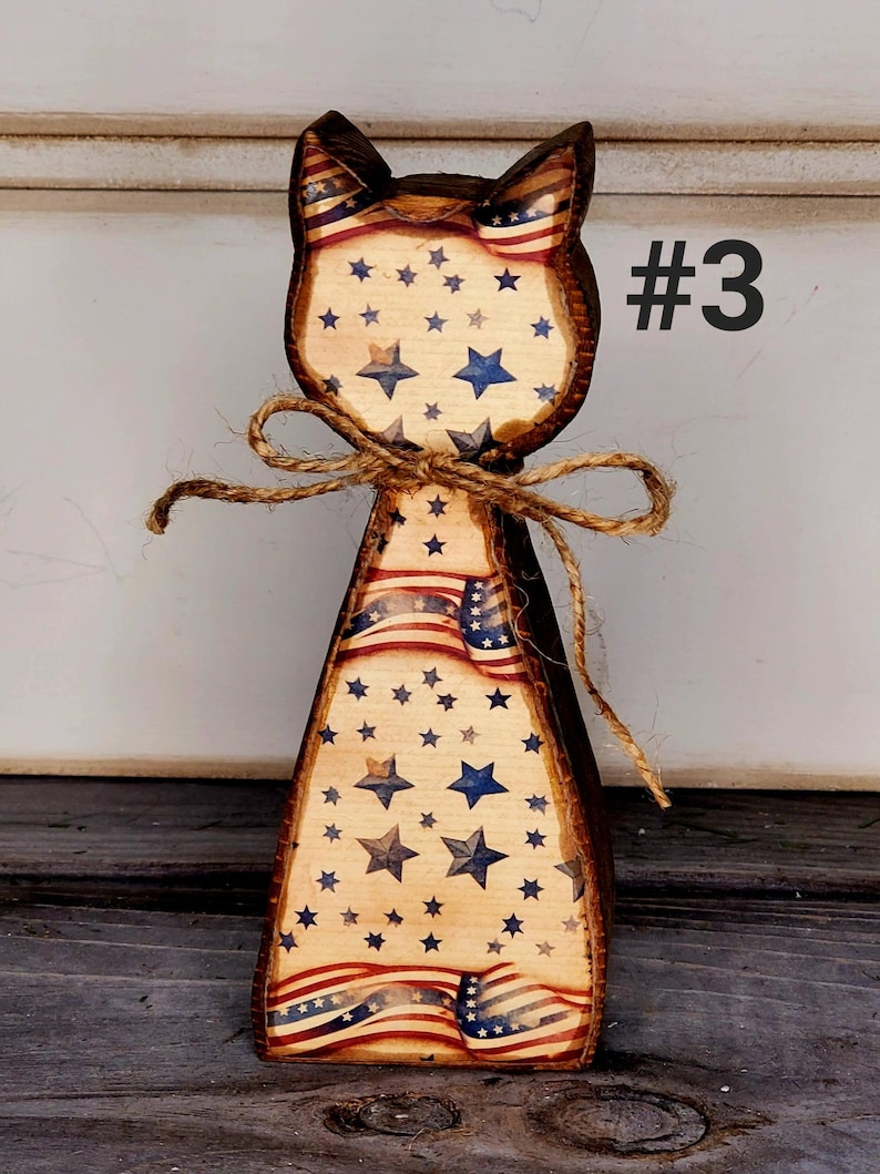 May include: Wooden cat-shaped decoration with a patriotic theme. The cat features a brown outline, with a design of the American flag and blue stars. A twine bow is tied around the neck. The number "#3" is printed on the right side.