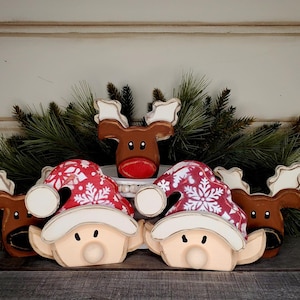 May include: Wooden Christmas decorations featuring elf and reindeer figures. The elves wear red hats with white snowflake patterns and white trim. The reindeer have brown bodies and antlers. Green pine branches are in the background.