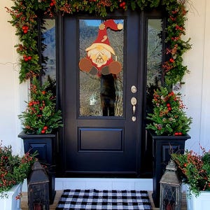 May include: A black front door with a Christmas wreath and a gnome decoration. Two black planters with greenery and red berries flank the door. A black and white checkered doormat sits in front of the door.