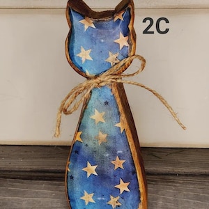 May include: Wooden cat figurine with a blue body adorned with gold stars. The cat has a brown outline and ears, and a twine bow around its neck. The number "2C" is visible on the right side of the cat.