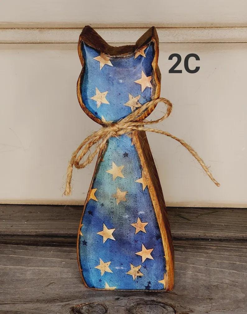 May include: Wooden cat figurine with a blue body adorned with gold stars. The cat has a brown outline and ears, and a twine bow around its neck. The number "2C" is visible on the right side of the cat.