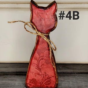 May include: A wooden cat-shaped decoration with a red floral design. The cat has a brown outline and is tied with twine. The text "#4B" is visible on the right side of the cat.