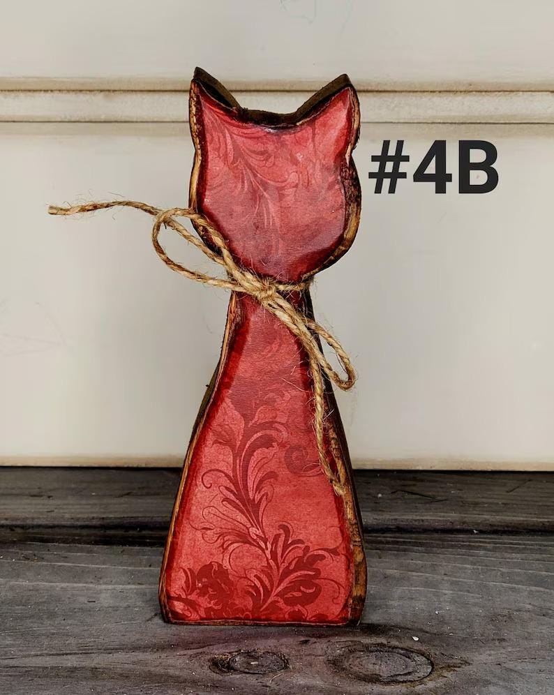 May include: A wooden cat-shaped decoration with a red floral design. The cat has a brown outline and is tied with twine. The text "#4B" is visible on the right side of the cat.