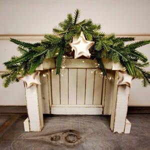 May include: A white wooden nativity stable structure featuring a distressed finish, adorned with green pine branches across the top. Three off-white star ornaments are attached to the front, and small warm white string lights are woven throughout.