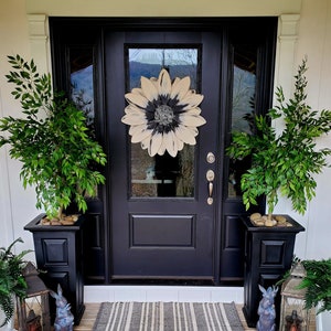 Könnte beinhalten: Eine schwarze Haustür mit einem weißen und schwarzen Sonnenblumenkranz. Zwei Topfpflanzen mit grünen Blättern stehen auf beiden Seiten der Tür. Eine gestreifte Fußmatte liegt auf der Veranda.
