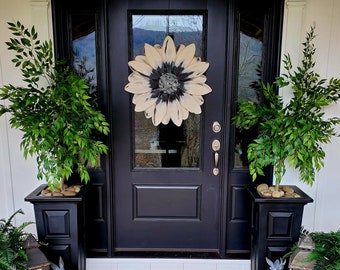 May include: A black front door with a white and black sunflower wreath. Two potted plants with green leaves are on either side of the door. A striped doormat is on the porch.