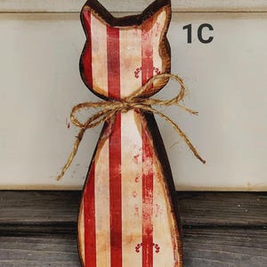 May include: Wooden cat-shaped decoration with red and white vertical stripes. The cat has a brown outline and is tied with a twine bow. The number "1C" is visible on the right side. The cat is standing on a wooden surface.