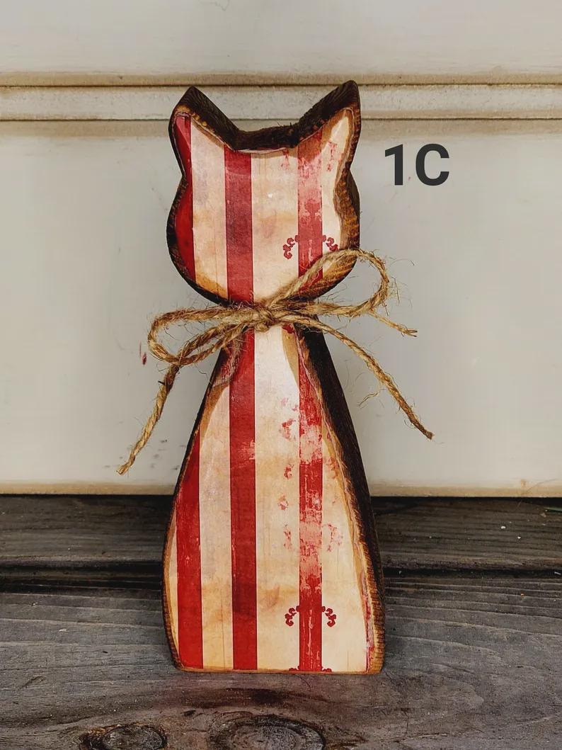 May include: Wooden cat-shaped decoration with red and white vertical stripes. The cat has a brown outline and is tied with a twine bow. The number "1C" is visible on the right side. The cat is standing on a wooden surface.