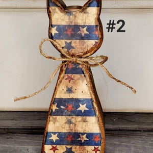 May include: Wooden cat-shaped decoration with a patriotic design. The cat has a distressed finish with blue and red stripes and stars. A twine bow is tied around the neck. The number "#2" is printed on the side.