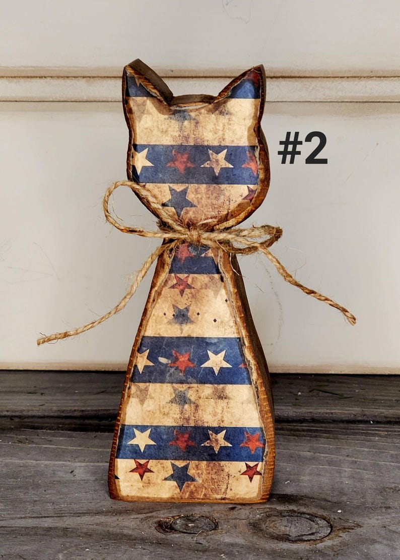 May include: Wooden cat-shaped decoration with a patriotic design. The cat has a distressed finish with blue and red stripes and stars. A twine bow is tied around the neck. The number "#2" is printed on the side.