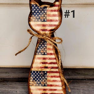 May include: Wooden cat-shaped decoration with a distressed American flag design. The cat has a rustic appearance, with a jute twine bow around its neck. The number "#1" is printed in black on the right side.