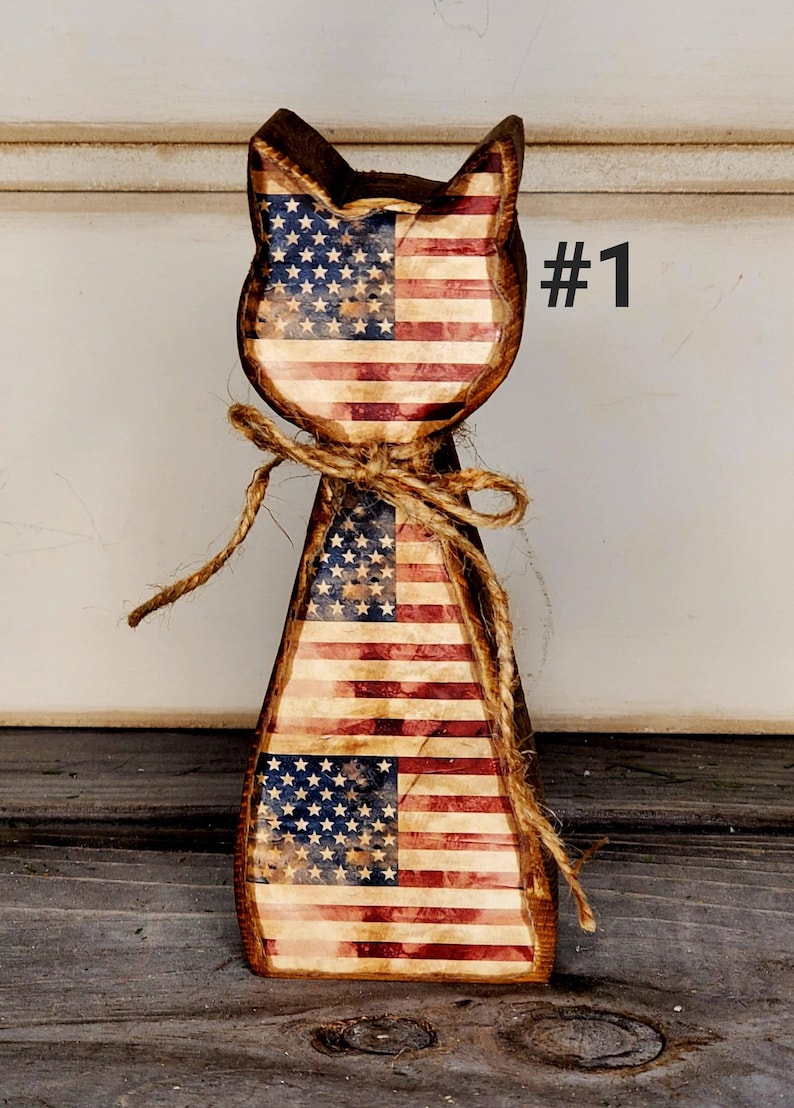 May include: Wooden cat-shaped decoration with a distressed American flag design. The cat has a rustic appearance, with a jute twine bow around its neck. The number "#1" is printed in black on the right side.