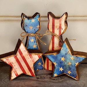 May include: Four wooden patriotic decorations. Two cat-shaped figures with blue star-covered faces and red and white striped bodies. Two star-shaped figures with red and white stripes and blue star-covered faces. All are tied with twine.