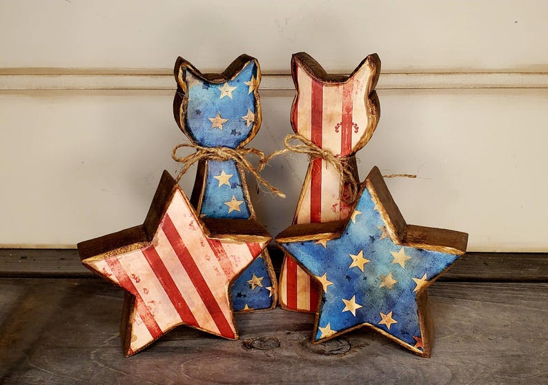 May include: Four wooden patriotic decorations. Two cat-shaped figures with blue star-covered faces and red and white striped bodies. Two star-shaped figures with red and white stripes and blue star-covered faces. All are tied with twine.