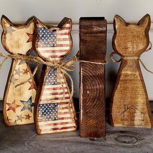 May include: Wooden cat figurines with patriotic American flag and star designs. Two cats feature the flag, while the others have star patterns. All are tied with twine. A dark brown wooden block is also present.