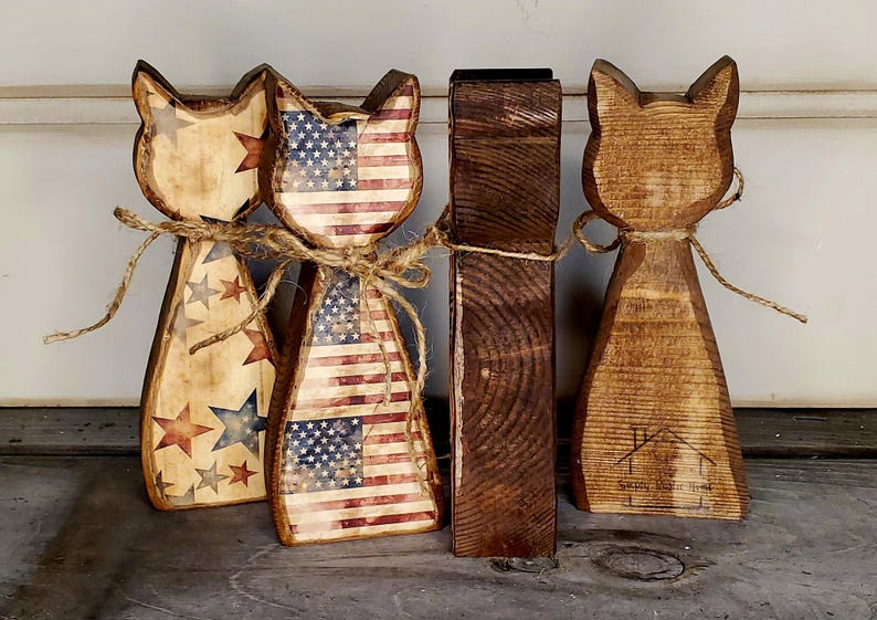 May include: Wooden cat figurines with patriotic American flag and star designs. Two cats feature the flag, while the others have star patterns. All are tied with twine. A dark brown wooden block is also present.