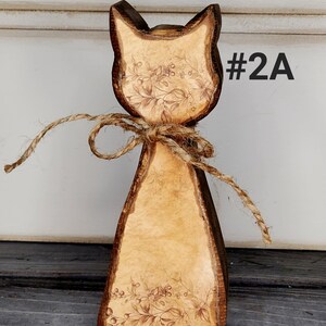 May include: Wooden cat figurine with a rustic design. The cat has a light brown body with a floral pattern and dark brown edges. A twine bow is tied around the neck. The text "#2A" is visible on the right side.