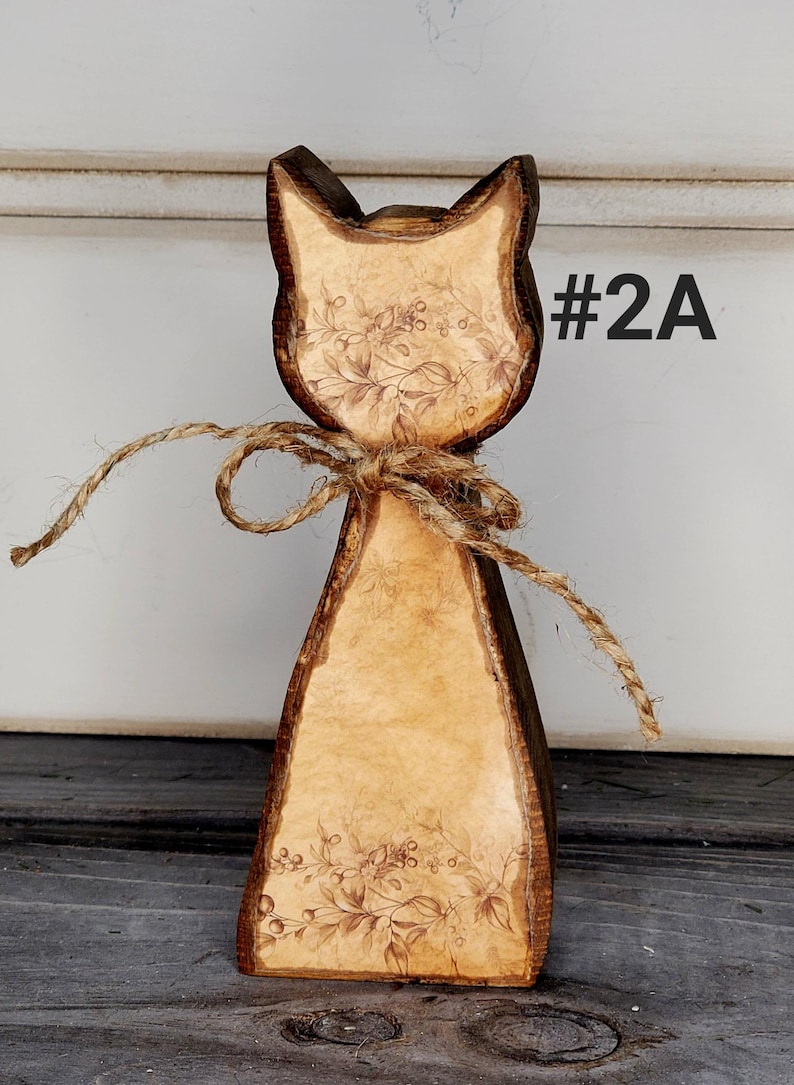 May include: Wooden cat figurine with a rustic design. The cat has a light brown body with a floral pattern and dark brown edges. A twine bow is tied around the neck. The text "#2A" is visible on the right side.