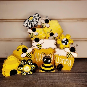 May include: A collection of wooden decorative items featuring yellow and black bees, yellow honeycombs, a yellow beehive, yellow flowers, and two yellow jars with the word Honey written in white script. The set includes various bee-themed shapes.