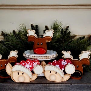 May include: A festive holiday display featuring wooden reindeer and elf-like figures. The reindeer have brown bodies, white antlers, and red noses. The elves have tan faces and white hats. Red and white snowflake-patterned hearts are also included.