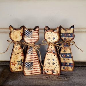 May include: Four wooden cat figurines with patriotic American flag and star designs. Each cat is tied with twine. The cats are painted with red, white, and blue colors, and are a decorative item.
