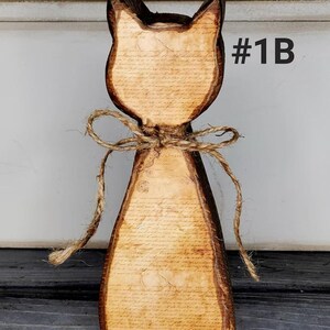 May include: Wooden cat-shaped figurine with a rustic design. The cat has a light brown body with a printed script pattern and dark brown edges. A twine bow adorns the neck. The text "#1B" is visible.