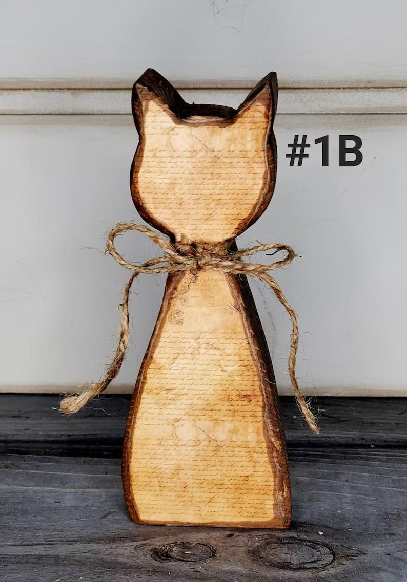 May include: Wooden cat-shaped figurine with a rustic design. The cat has a light brown body with a printed script pattern and dark brown edges. A twine bow adorns the neck. The text "#1B" is visible.