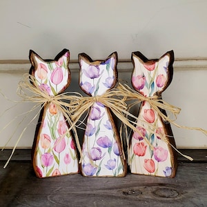 May include: Three wooden cat figurines with a floral tulip design in pink, purple, and white. Each cat has a dark brown outline and is adorned with a raffia bow around the neck. The cats are standing on a wooden surface.