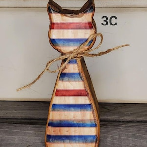 May include: Wooden cat figurine with a patriotic red, white, and blue striped design. The cat has a rustic, hand-crafted appearance, with a twine bow around its neck. The number "3C" is visible on the right side.