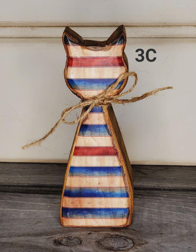 May include: Wooden cat figurine with a patriotic red, white, and blue striped design. The cat has a rustic, hand-crafted appearance, with a twine bow around its neck. The number "3C" is visible on the right side.