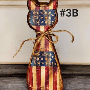 May include: Wooden cat figurine with a distressed American flag design. The cat has a rustic, vintage look with red, white, and blue colors. A twine bow is tied around the neck. The text "#3B" is visible on the right side.