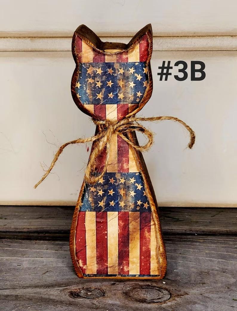 May include: Wooden cat figurine with a distressed American flag design. The cat has a rustic, vintage look with red, white, and blue colors. A twine bow is tied around the neck. The text "#3B" is visible on the right side.