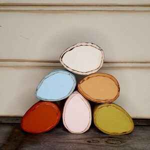 May include: A pyramid of six painted wooden egg shapes in various colors: white, light blue, orange, pink, tan, and green. Each egg has a dark brown outline. The eggs are arranged on a dark wooden surface.