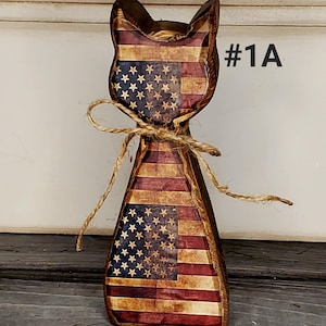May include: Wooden cat figurine with an American flag design. The cat-shaped decoration features red and white stripes and blue sections with white stars. A twine bow is tied around the neck. The text "#1A" is visible.