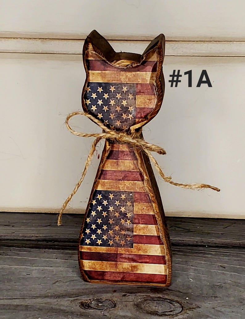 May include: Wooden cat figurine with an American flag design. The cat-shaped decoration features red and white stripes and blue sections with white stars. A twine bow is tied around the neck. The text "#1A" is visible.