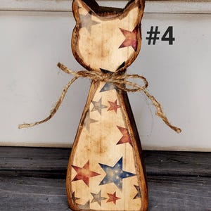 May include: Wooden cat figurine with a rustic finish, adorned with red, blue, and gray stars. The cat is tied with twine and features the number "#4" in black. The figurine has a distressed, vintage appearance.