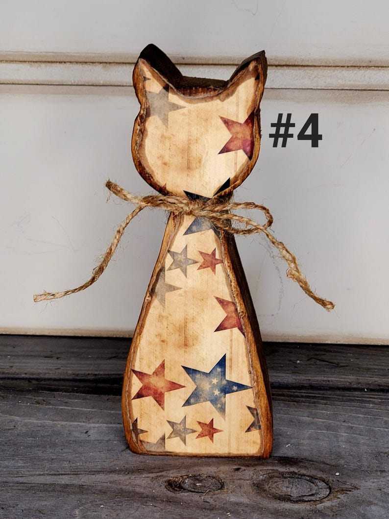 May include: Wooden cat figurine with a rustic finish, adorned with red, blue, and gray stars. The cat is tied with twine and features the number "#4" in black. The figurine has a distressed, vintage appearance.