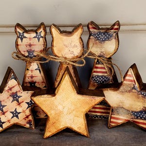 May include: A collection of wooden patriotic decorations, including star and cat-shaped figures. The stars and cat figures are painted with a distressed American flag design in red, white, and blue. The figures are tied together with twine.