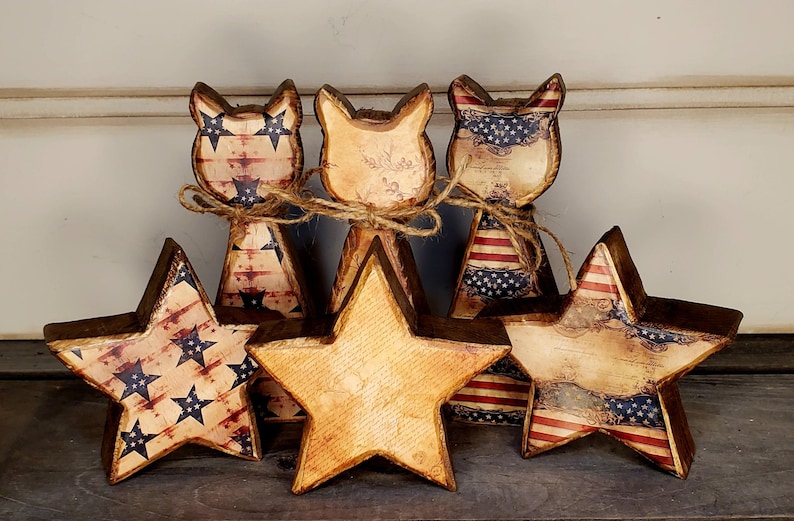 May include: A collection of wooden patriotic decorations, including star and cat-shaped figures. The stars and cat figures are painted with a distressed American flag design in red, white, and blue. The figures are tied together with twine.
