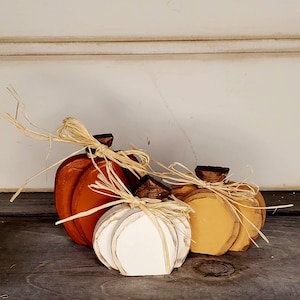 May include: Three wooden pumpkin-shaped decorations in orange, white, and yellow. Each pumpkin features a dark brown wooden stem and is tied with a natural raffia bow. The pumpkins are arranged in a cluster with the orange one in the back.