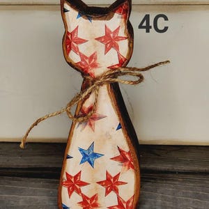 May include: Wooden cat figurine with a patriotic design. The cat is painted with red and blue stars on a cream background, with a brown outline. A twine bow is tied around the neck. The number "4C" is visible on the right side.