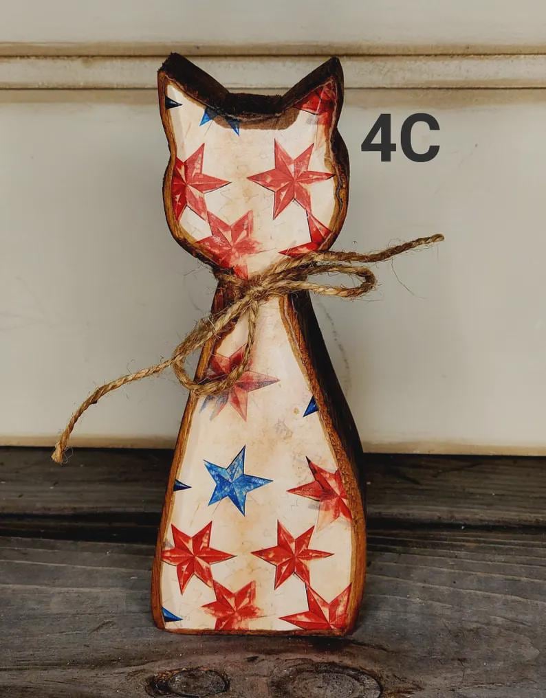 May include: Wooden cat figurine with a patriotic design. The cat is painted with red and blue stars on a cream background, with a brown outline. A twine bow is tied around the neck. The number "4C" is visible on the right side.