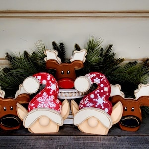 May include: Christmas-themed wooden decorations featuring reindeer and gnome figures. The reindeer are brown with white antlers, and the gnomes wear red hats with white snowflake patterns. Green pine branches are in the background.