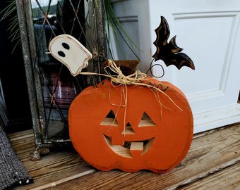 Könnte beinhalten: Eine Halloween-Kürbisdekoration aus Holz mit geschnitztem Gesicht, orangefarbener Farbe und einer Strohschleife. Ein kleiner Holzgeist und eine Fledermaus sind befestigt. Der Kürbis steht auf einer Holzoberfläche.