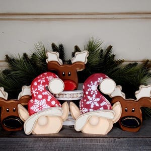 May include: Wooden Christmas decorations featuring reindeer and gnomes. The reindeer are brown with white antlers, and the gnomes wear red hats with white snowflake patterns. Green pine branches are in the background.