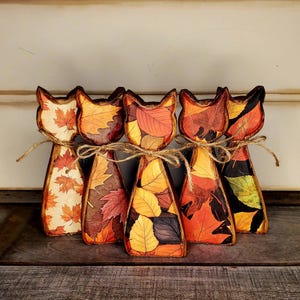 Op de afbeelding: Vijf houten katvormige beeldjes versierd met diverse herfstbladpatronen in tinten oranje, rood, geel en bruin. Elk katfiguur is voorzien van een strik van natuurlijk touw om de nek en toont een uniek ontwerp met herfstbladeren.