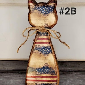 May include: Wooden cat-shaped decoration with an American flag design. The cat has a rustic appearance with a distressed finish, featuring red and white stripes and a blue field of stars. A twine bow is tied around the neck, and the text "#2B" is visible.