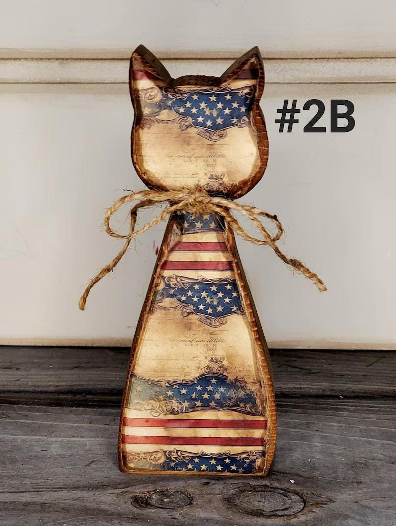 May include: Wooden cat-shaped decoration with an American flag design. The cat has a rustic appearance with a distressed finish, featuring red and white stripes and a blue field of stars. A twine bow is tied around the neck, and the text "#2B" is visible.