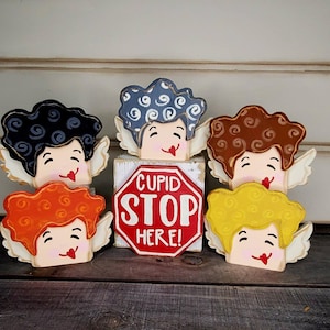 May include: Wooden cherub figurines with various hair colors, including black, orange, brown, blue, and yellow, each with white wings and a smiling face. A red stop sign with the text "Cupid Stop Here!" is in the center.